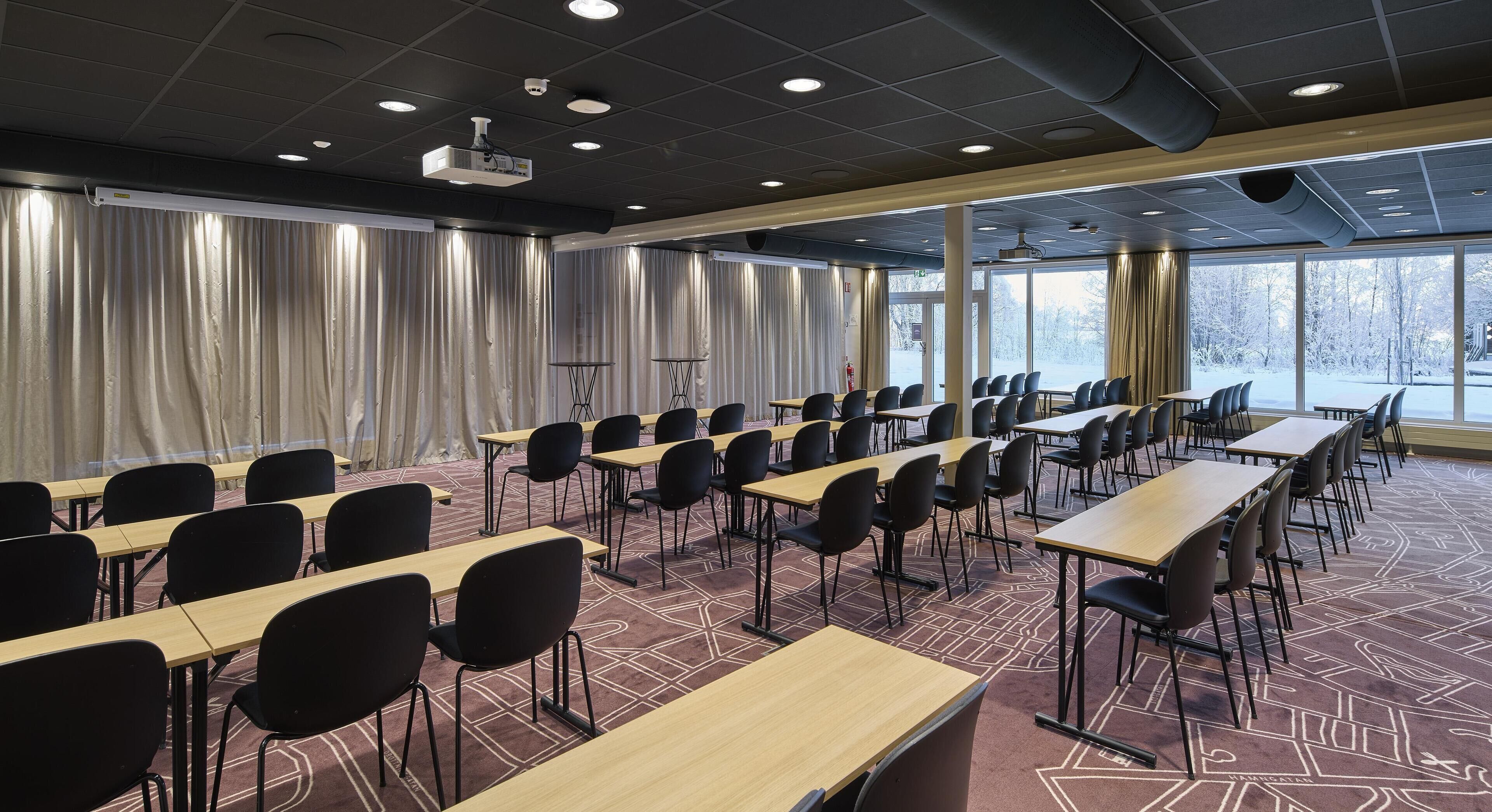 Un salón de conferencias con mesas y sillas, ventanas amplias, cortinas y proyectores de techo.