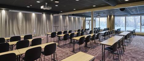 Un salón de conferencias con mesas y sillas, ventanas amplias, cortinas y proyectores de techo.