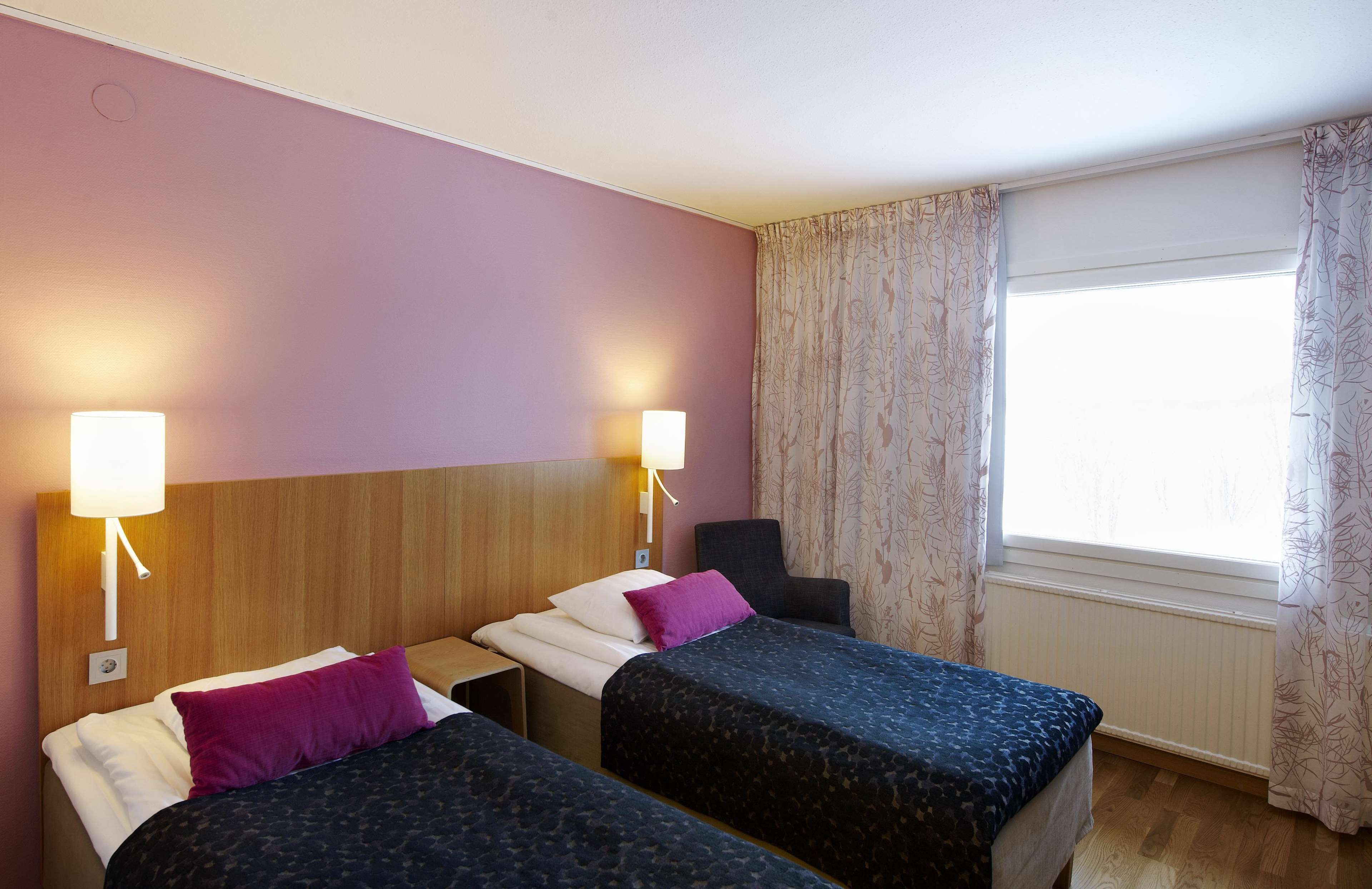 A hotel room with two beds, a wooden headboard, a nightstand, a chair, and a window with curtains.