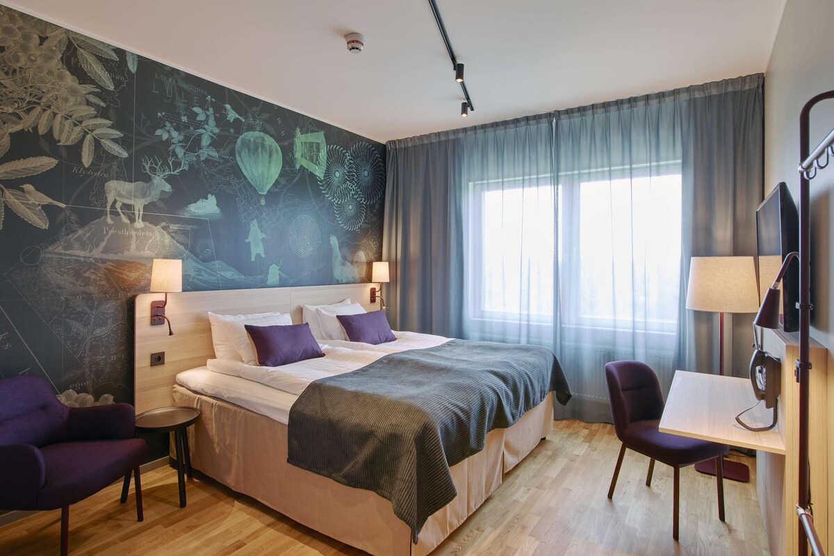Una habitación de hotel moderna con una cama grande, un escritorio, una silla y un mural en la pared.