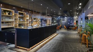 A modern bar area with a long counter, shelves with bottles and glasses, and a seating area with orange chairs. - Scandic Luleå (Luleå)