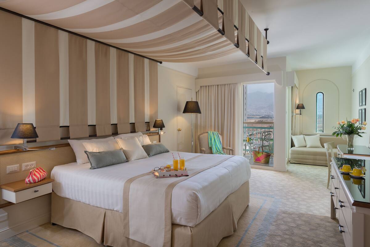 Ein Hotelzimmer mit einem großen Bett, Nachttischen, einem Beistelltisch, einem Sessel, einer Couch, einem Balkon mit Aussicht und einem Essbereich.