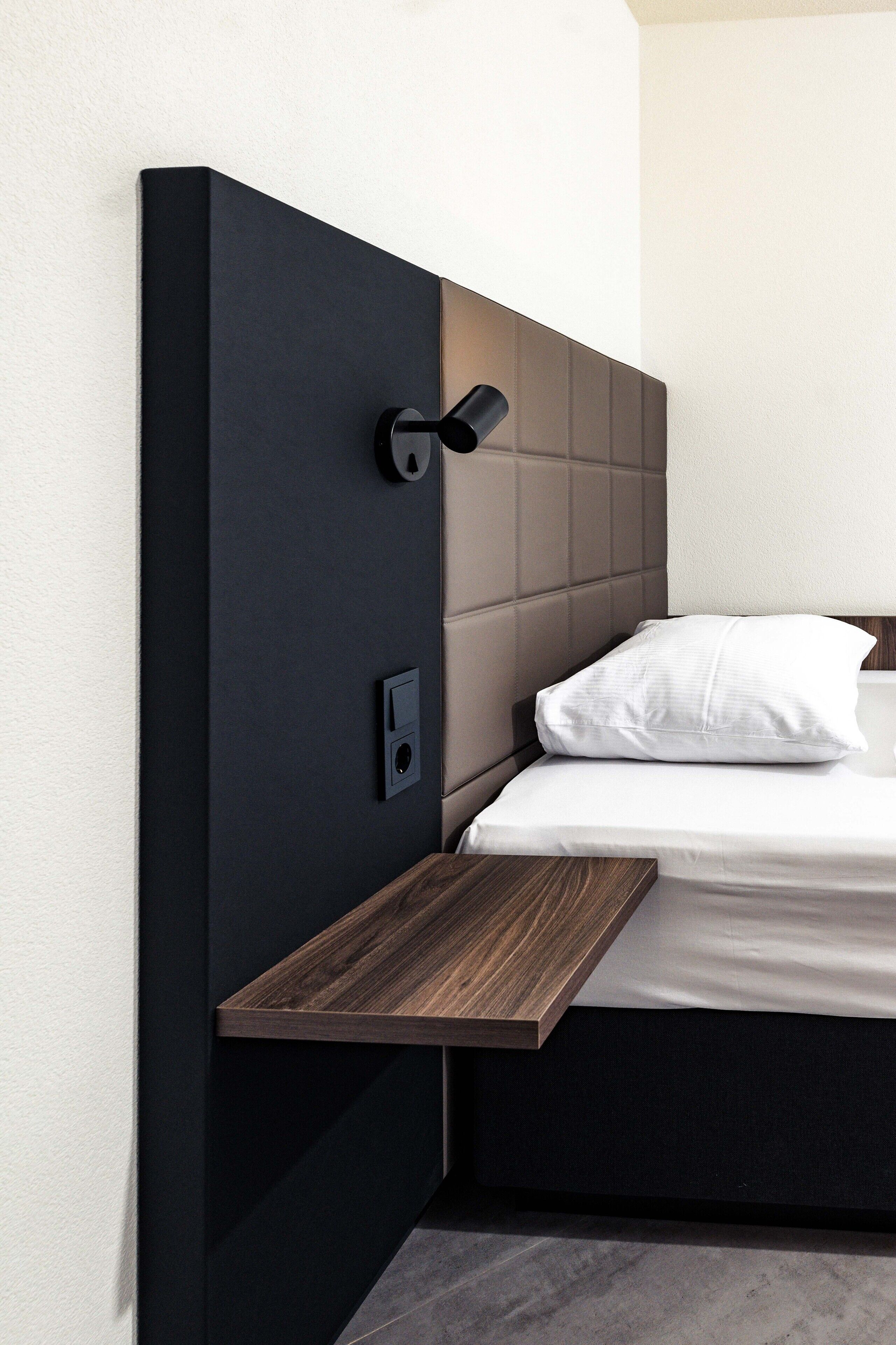 Una cama moderna con una mesita de noche de madera, cabecero con luz integrada y un pequeño panel de control.