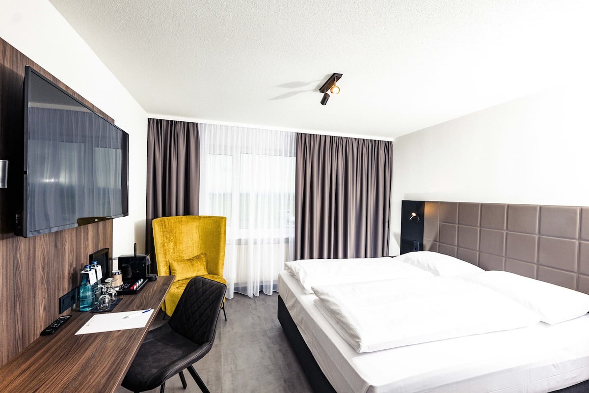 A hotel room with a bed, a desk with a chair, a TV, and a window with curtains.