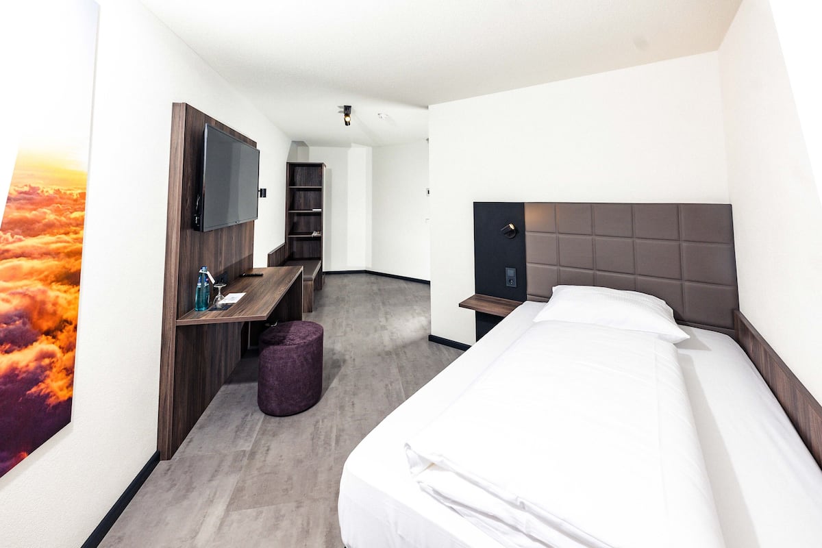 A hotel room with a bed, a desk with a TV, a bookshelf, and a large window with a sunset view.