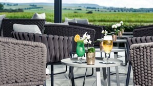 A patio area with wicker chairs and tables, two drinks on a table, and a view of green fields. - Sombea Donaueschingen (Donaueschingen)