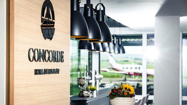A hotel reception area with a wooden wall displaying the name 'CONCORDIA HOTEL HUGSPAZ'. There are black pendant lights above a counter with a vase of flowers. - Sombea Donaueschingen (Donaueschingen)