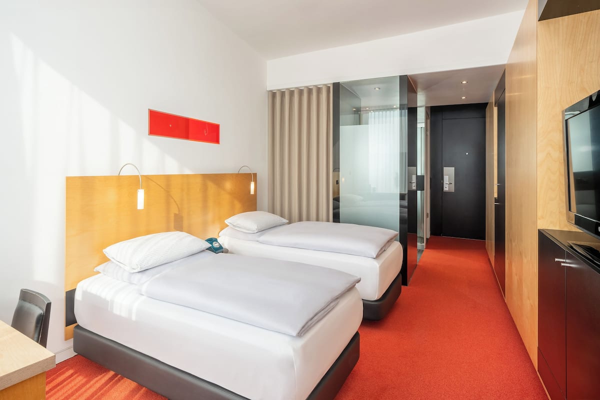 Une chambre d’hôtel avec deux lits, une tête de lit en bois, une moquette rouge, un téléviseur et une cloison en verre.