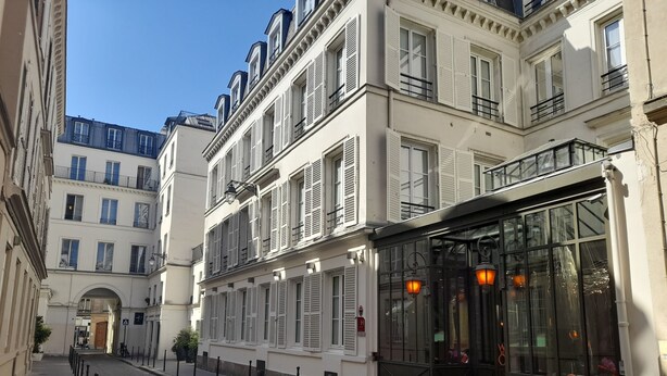 A cobblestone street lined with historic buildings, featuring a restaurant with large glass windows and outdoor seating. - WHO - Windsor Hotel Opera (Paris)