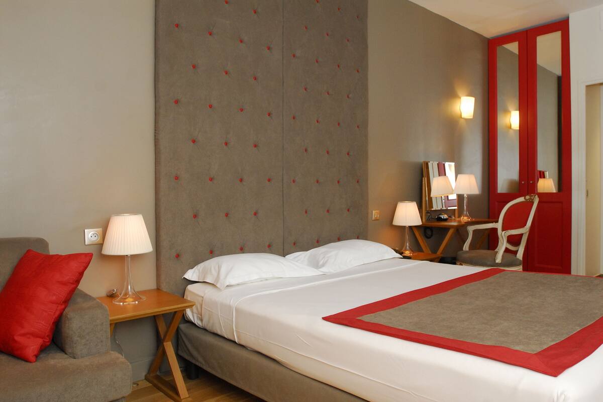 A hotel room with a bed, a nightstand with a lamp, a chair, and a desk with a chair.
