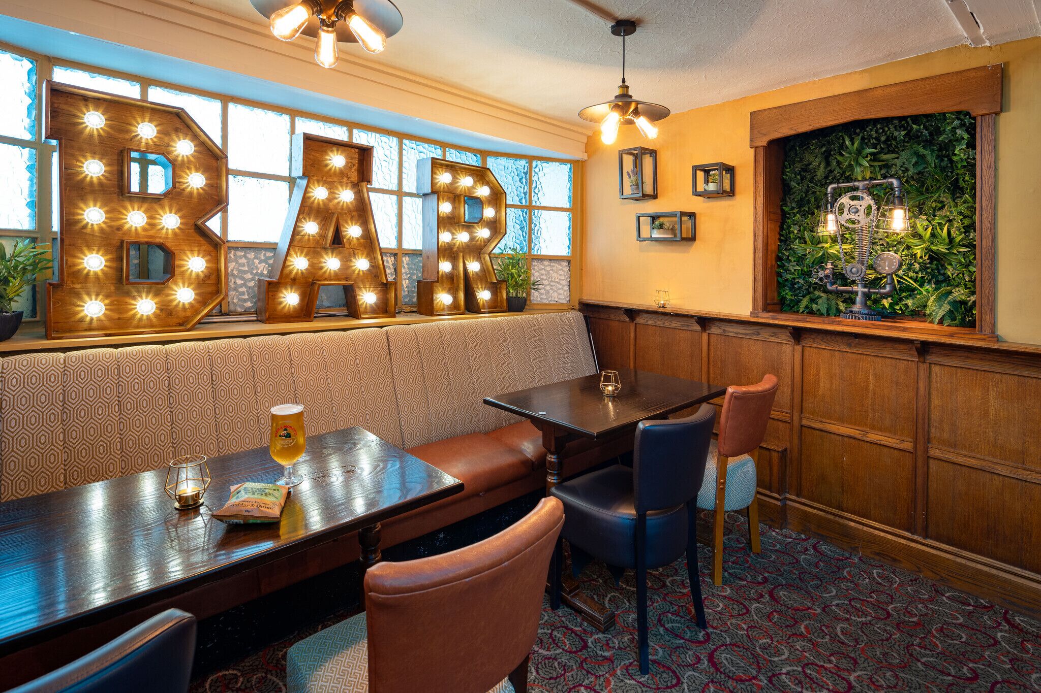 A bar area with illuminated letters spelling "BAR," a wooden bench, a table with a glass of beer, and a patterned carpet.