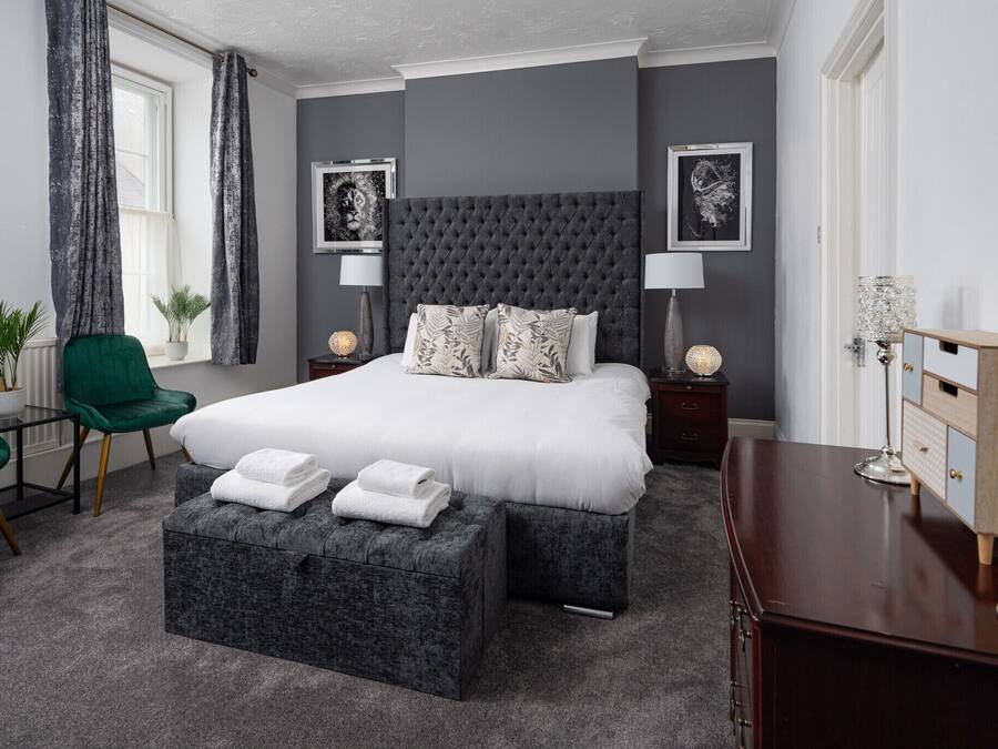 Une chambre à coucher avec un grand lit, deux tables de chevet, un fauteuil vert, une commode en bois et une fenêtre avec des rideaux.