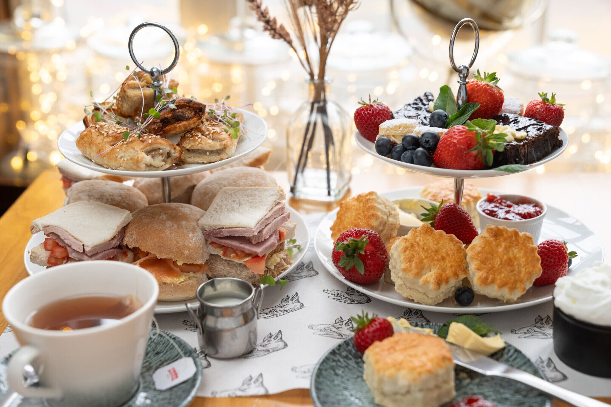 Una mesa con diferentes alimentos, incluyendo sándwiches, scones y pasteles, junto con tazas de té y una pequeña taza de metal.