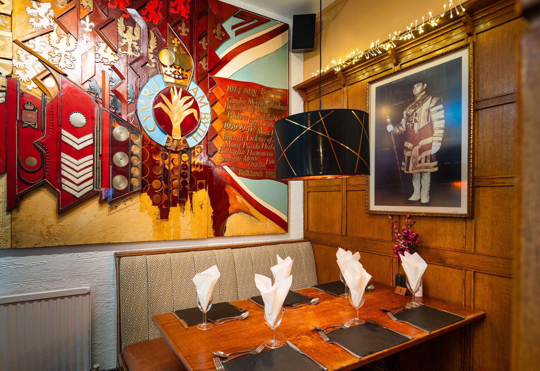 A dining area with a wooden table set for four, a decorative wall with a coat of arms, and a framed portrait.