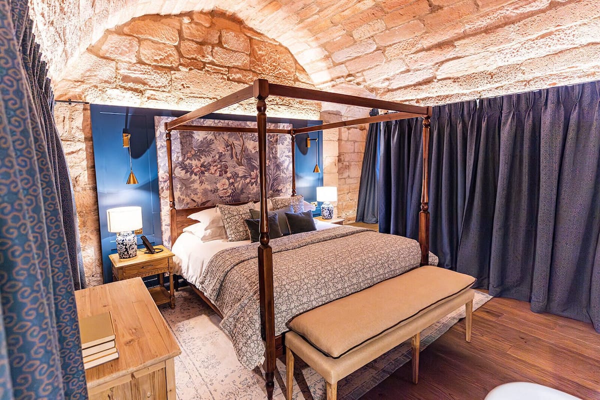 A four-poster bed in a room with stone walls, a wooden bench, a nightstand with a lamp, and a patterned wall.