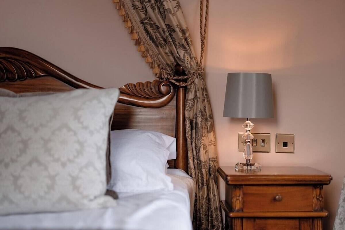 A neatly made bed with a headboard, a bedside table with a lamp, and a power outlet on the wall.