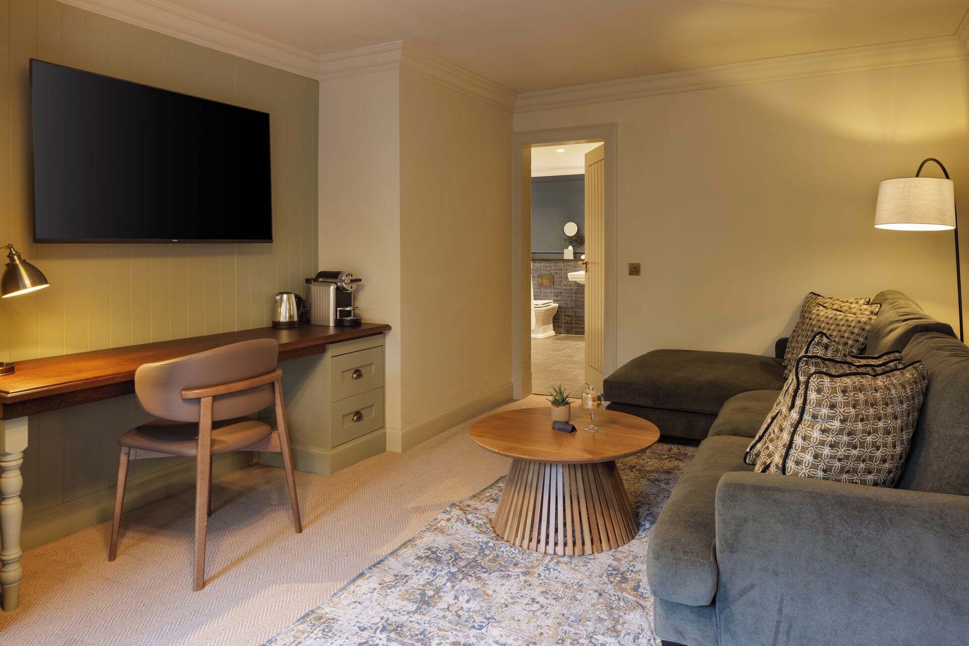A compact living space with a flat-screen TV, a wooden desk with a chair, a round coffee table, and a grey sofa with patterned cushions.