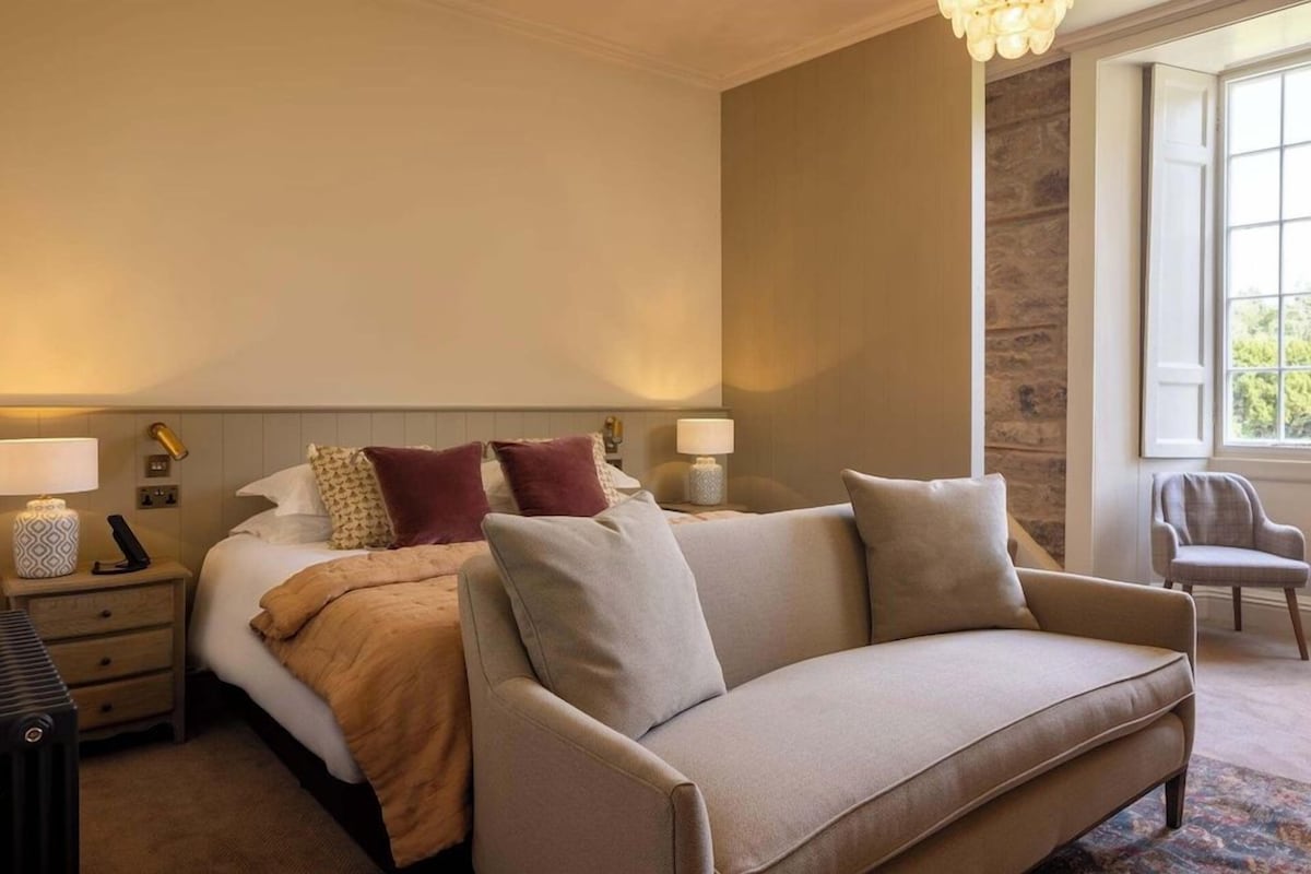 A hotel room with a bed, a sofa, a nightstand, a lamp, and a window.