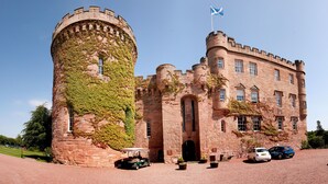 A castle with a round tower and a flag, surrounded by a gravel driveway and parked cars.
