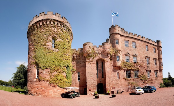 A castle with a round tower and a flag, surrounded by a gravel driveway and parked cars. - Dalhousie Castle (Bonnyrigg)