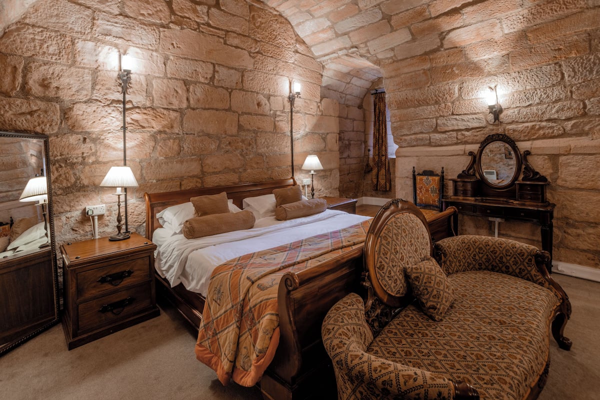 A room with a stone wall, a wooden bed, a chair, a dresser, and a mirror.