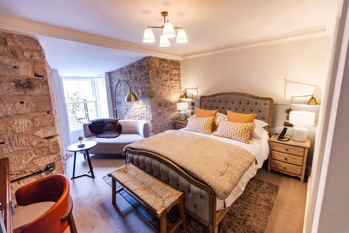 A bedroom with a stone wall, a bed with a wooden headboard, a bench, a small table, and a chair.