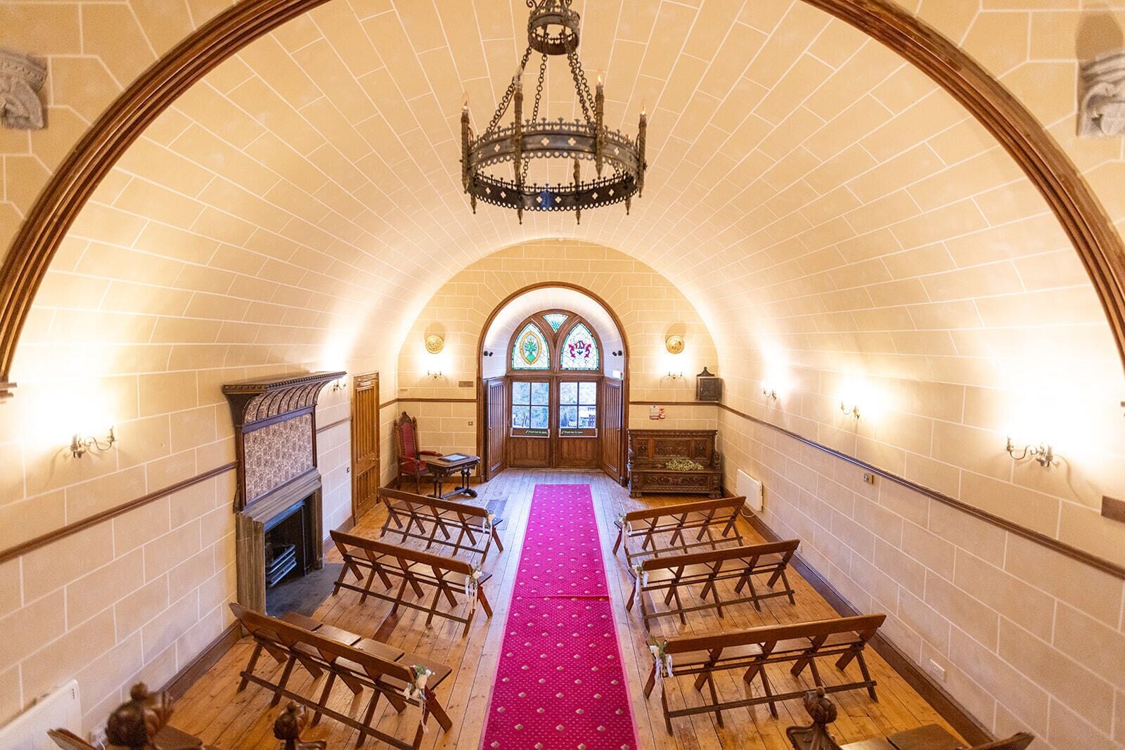 An arched interior with wooden pews, a central aisle, a chandelier, and a stained glass window.