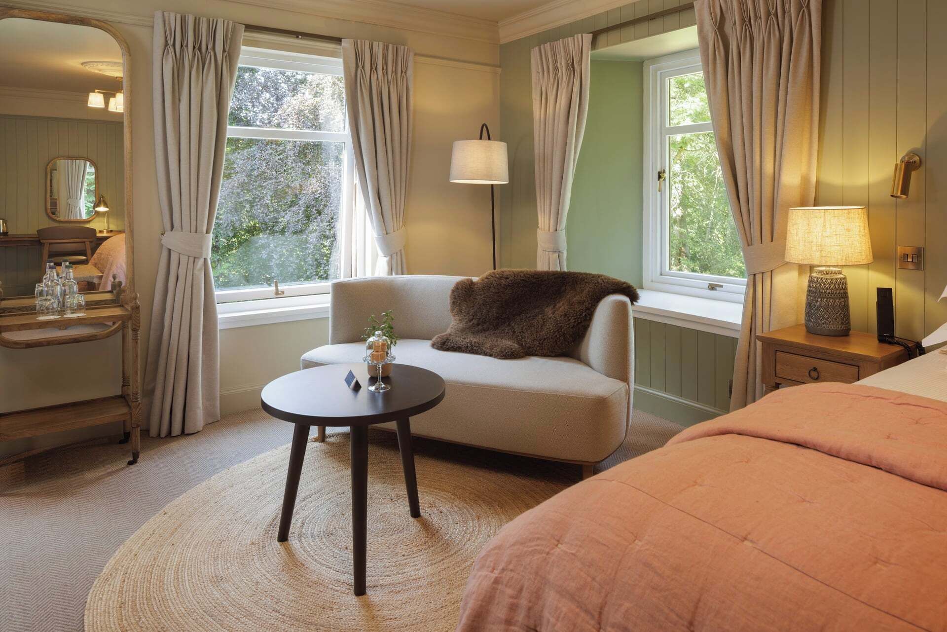Une chambre d’hôtel comprenant un lit, un canapé, une petite table, un miroir, une fenêtre avec des rideaux, une lampe et une table de chevet avec une lampe.