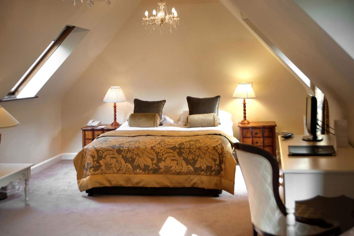A bedroom with a large bed, two bedside tables, a desk, a chair, a chandelier, and a skylight.