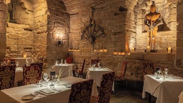 Interior de paredes de piedra con mesas dispuestas para comer, sillas con diseños y ambiente cálido a la luz de las velas.