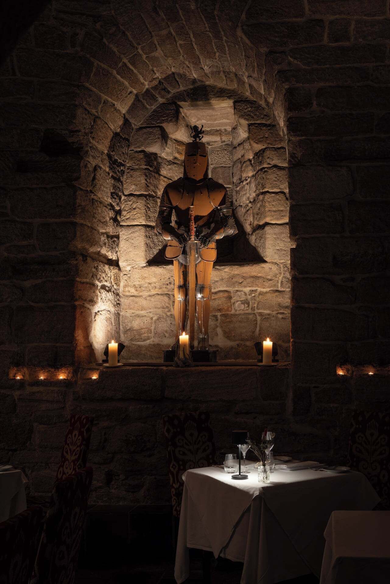 A stone wall with a statue, candles, and a table with glasses and a menu.