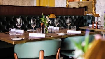 Un comedor con una mesa larga dispuesta para una comida, un asiento tipo banqueta de cuero negro al fondo y un jarrón con flores sobre la mesa.