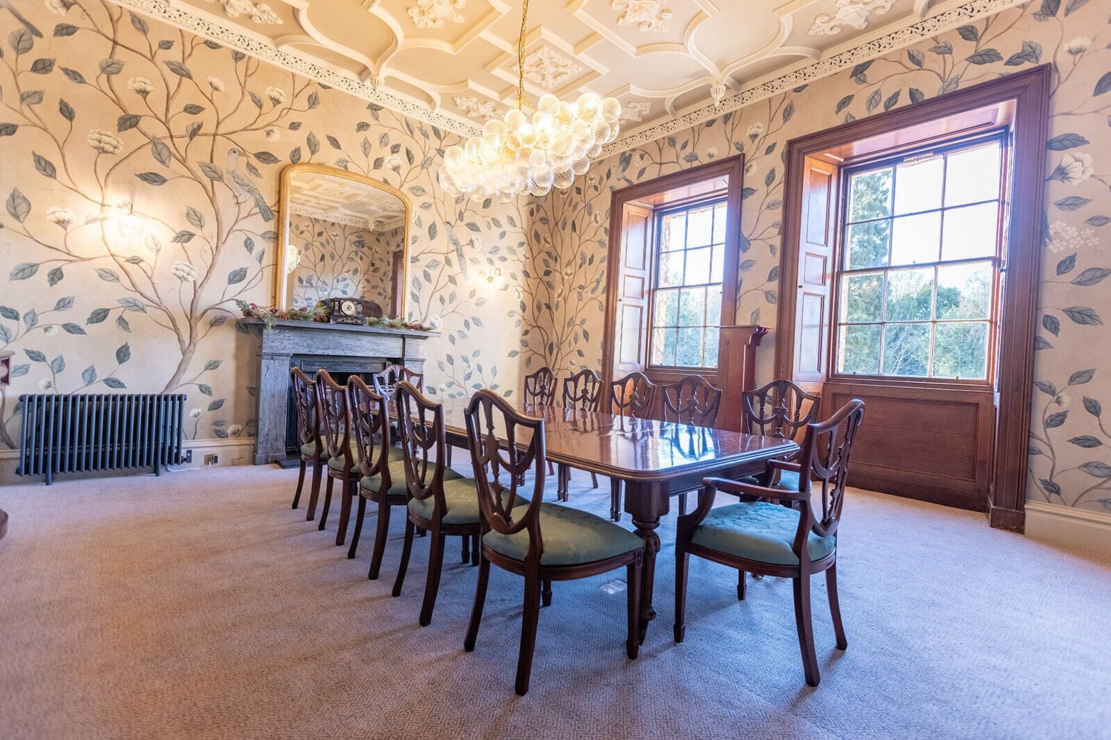 A dining room with a long table and chairs, a fireplace, and large windows.