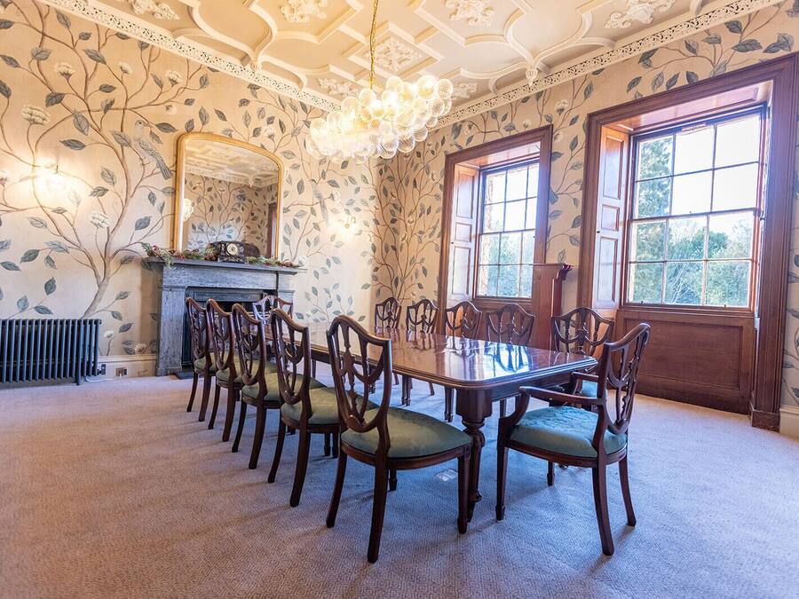 Een eetkamer met een lange tafel, stoelen, een open haard en grote ramen.