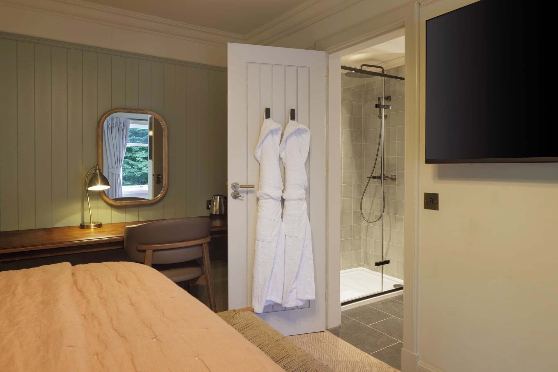 Una habitación de hotel con cama, escritorio con lámpara, bata colgada en la puerta, ducha y televisor montado en la pared.