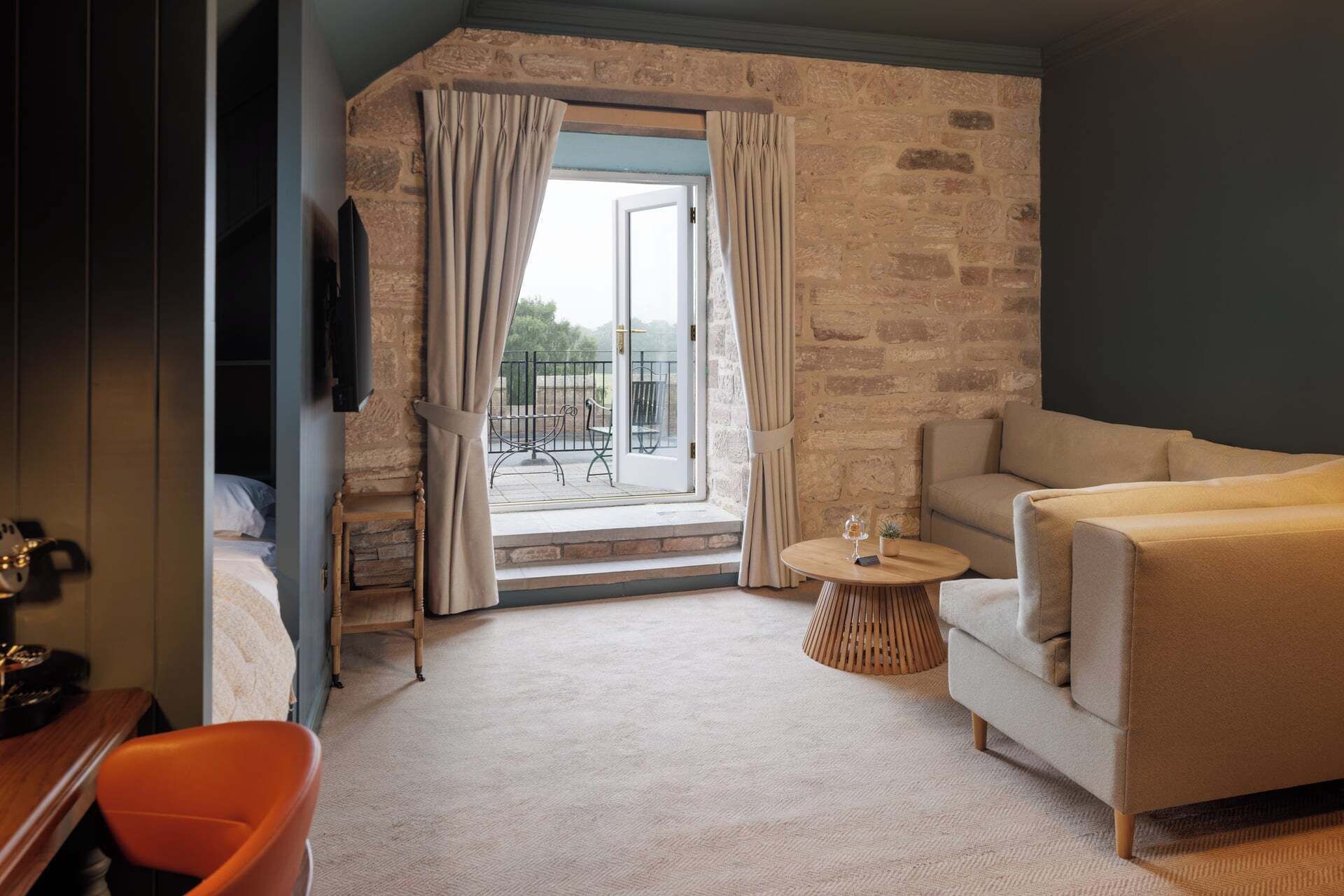A hotel room with a stone wall, a sofa, a small round table with a glass, and a view of the outdoors.
