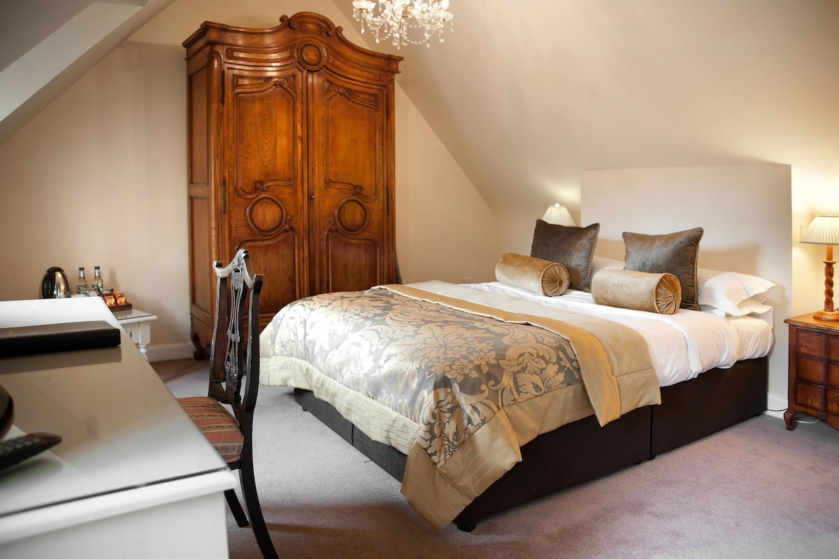 A bedroom with a large wooden wardrobe, a bed with a patterned bedspread, and a desk with a lamp.