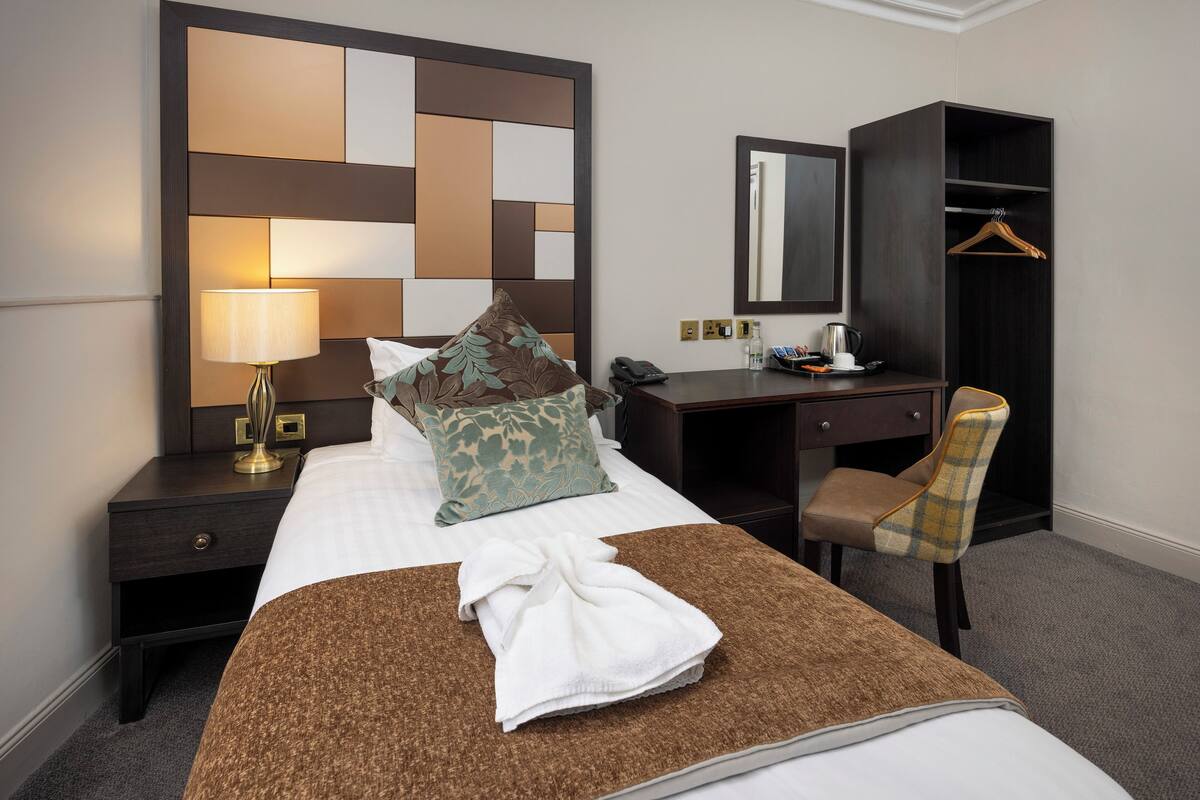 A hotel room with a bed, a desk, a chair, a wardrobe, a mirror, a lamp, and a towel on the bed.