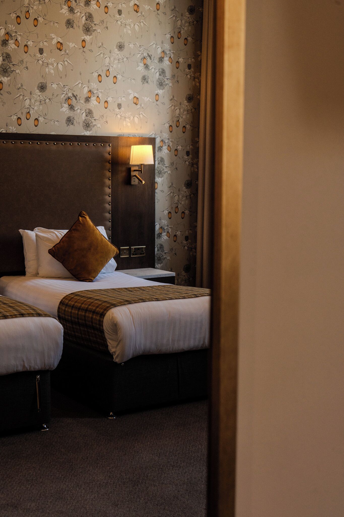 Ein Hotelzimmer mit zwei Betten, einem Nachttisch, einer Lampe und blumigem Wandpapier.