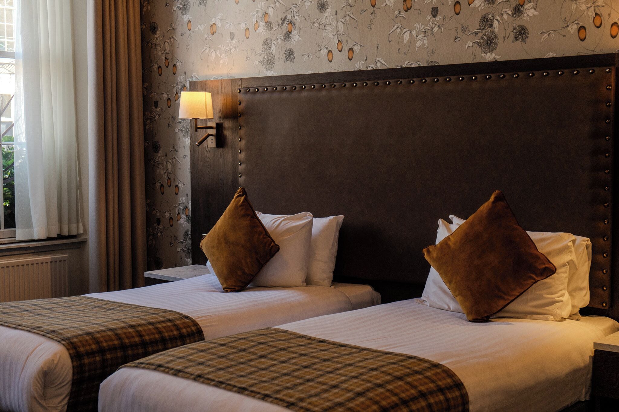 A hotel room with two beds, a headboard with a light fixture, and floral wallpaper.