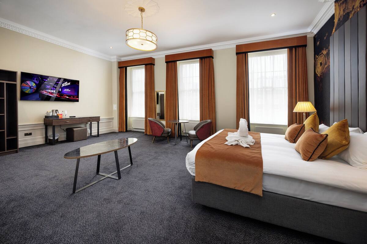A hotel room with a bed, desk, chairs, a television, and large windows.