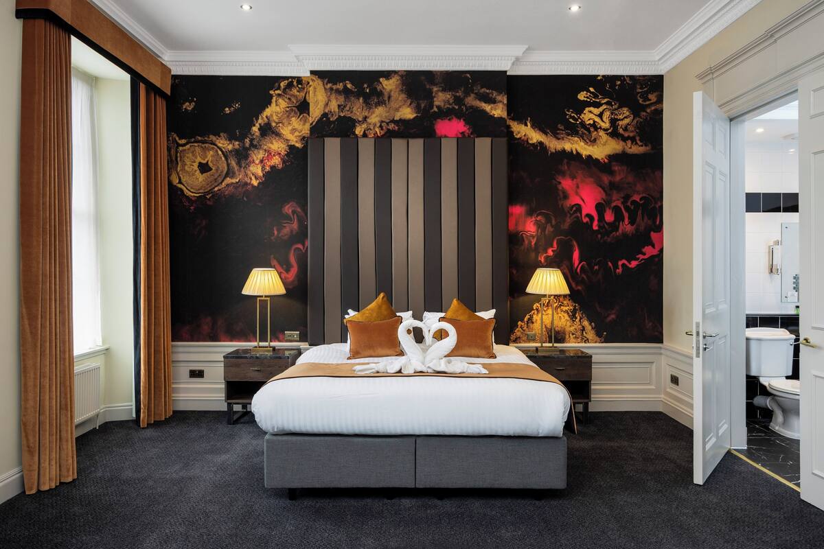 A hotel room with a large bed, two bedside tables with lamps, a dark carpet, and a mural on the wall.