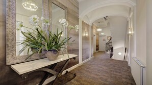 Un couloir avec un grand miroir, une plante en pot aux fleurs blanches, un lustre et un escalier.