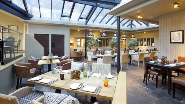 A spacious indoor area with a high ceiling and skylights, featuring multiple dining tables and chairs, and a kitchen area in the background. - The Broadway Hotel (Broadway)