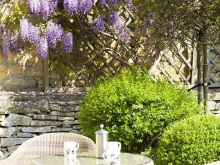 An outdoor seating area with wicker furniture and a table, surrounded by a stone wall and lush greenery.