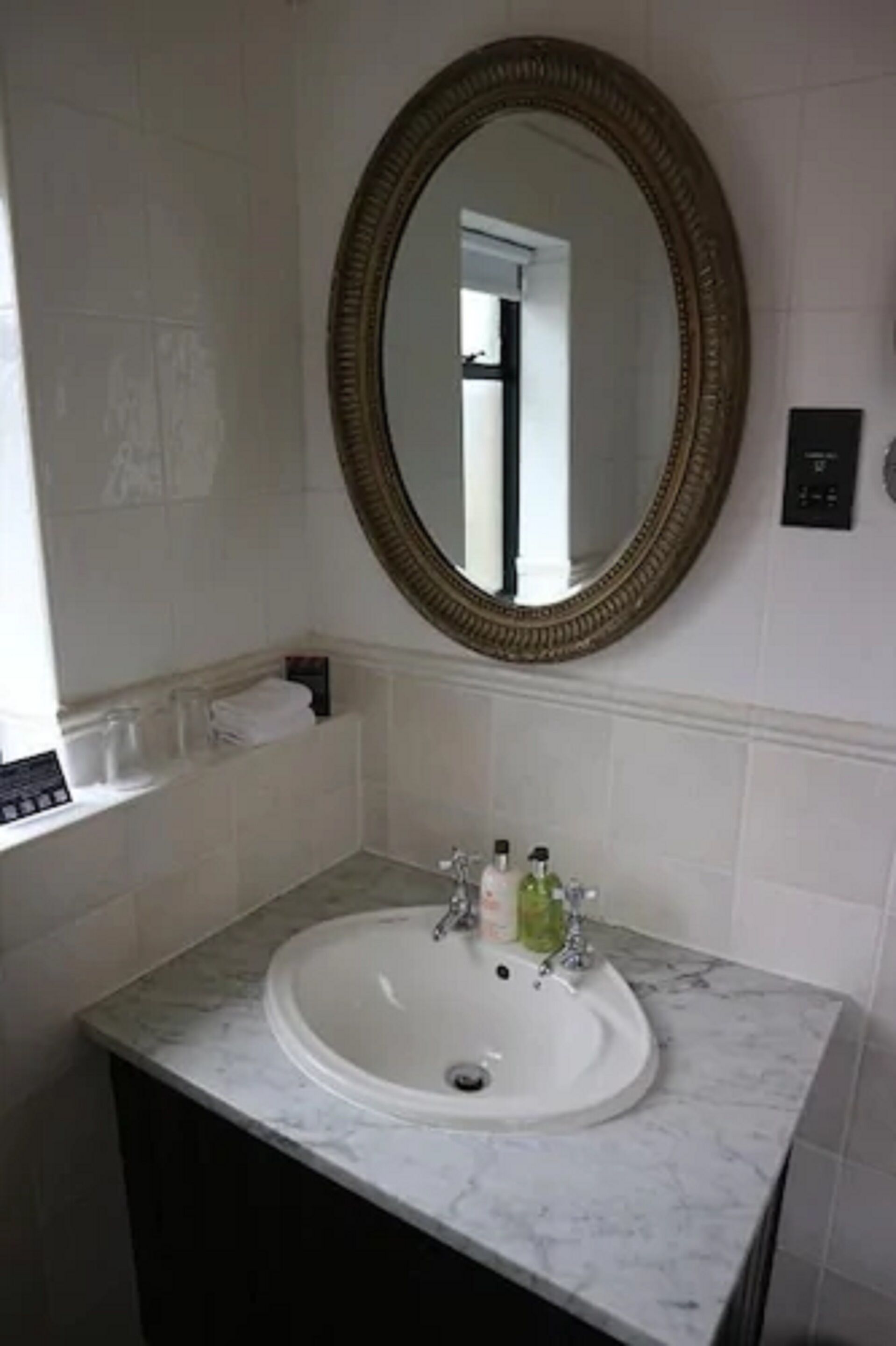 Un baño con un lavamanos blanco, un espejo con marco dorado y una ventana con vistas al interior.