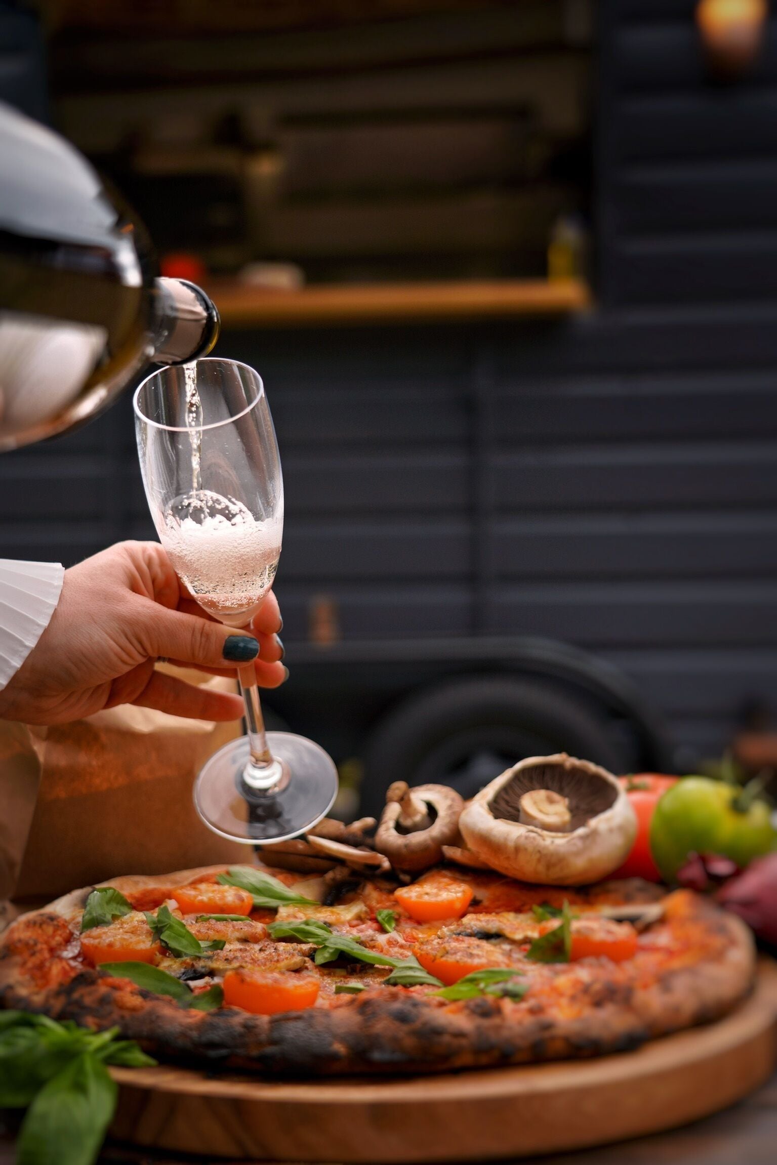 Una mano sosteniendo una copa de champán con burbujas, una pizza con tomates y albahaca, y champiñones sobre una tabla de madera.