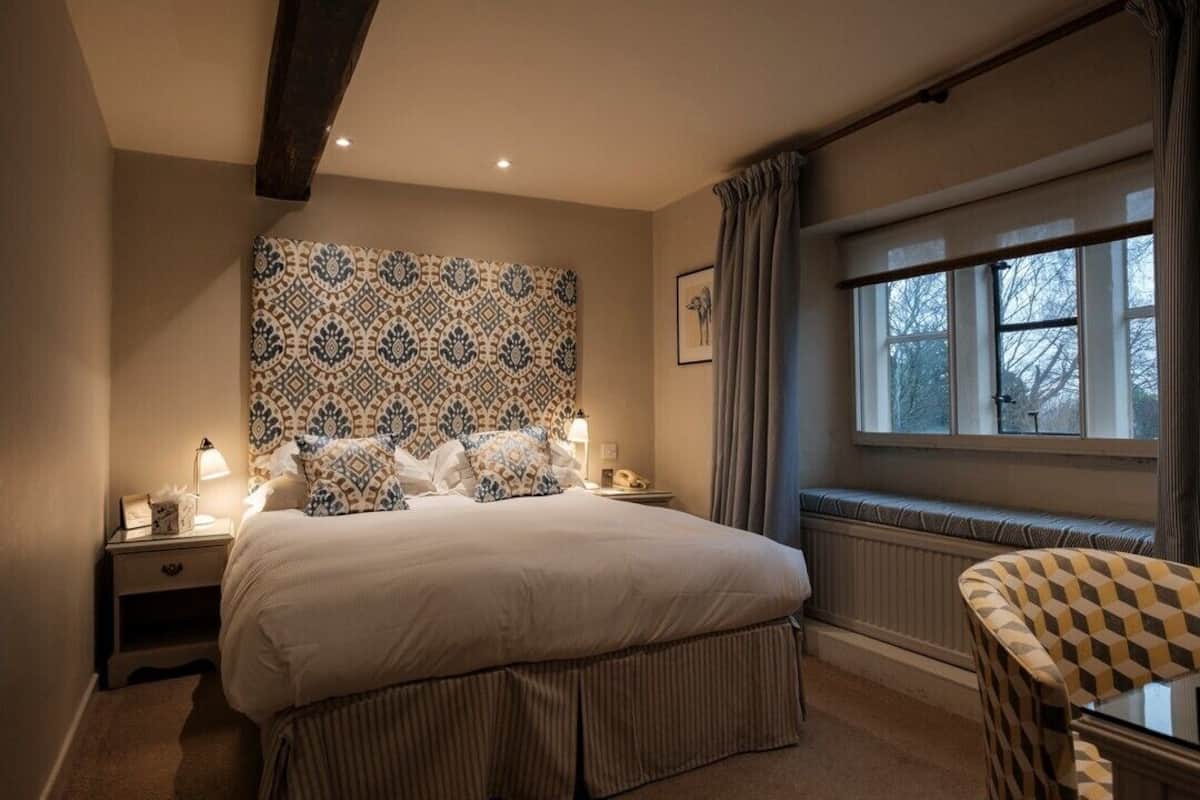A bedroom with a large bed, a patterned headboard, a nightstand with a lamp, a window with curtains, and a chair.