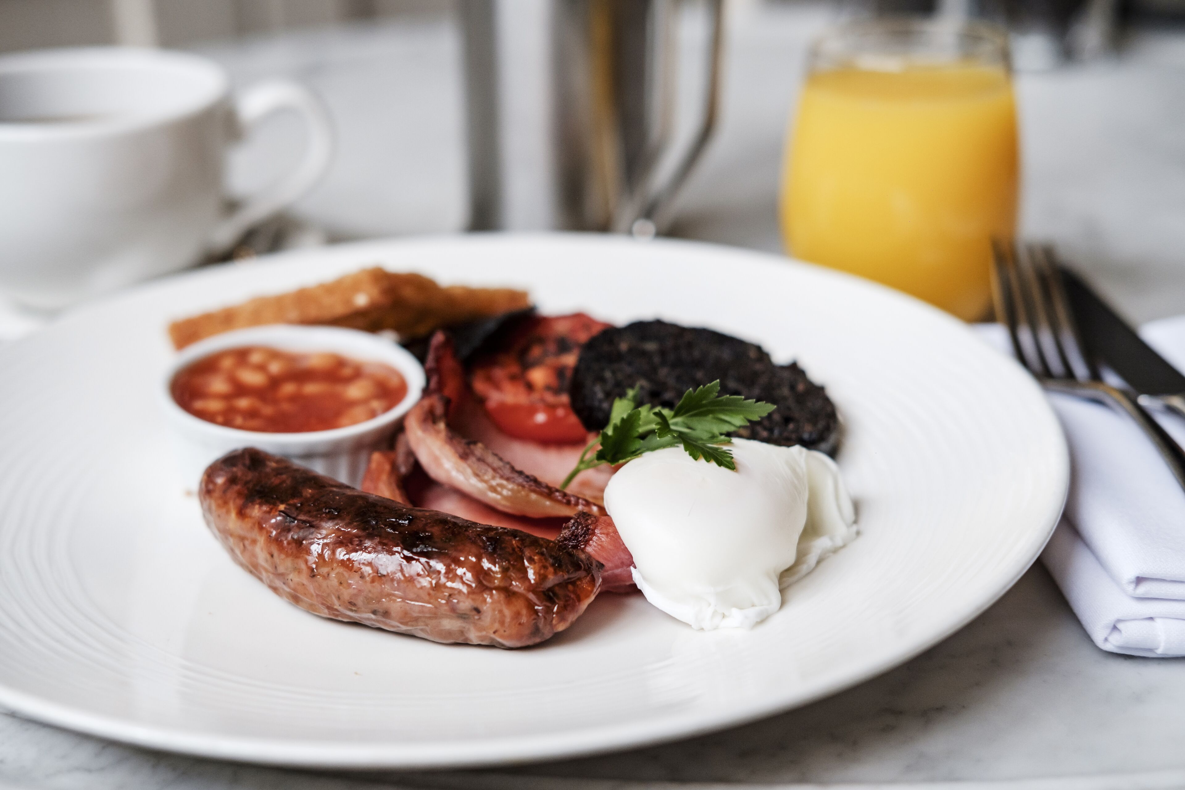 A plate of breakfast food including sausages, eggs, tomatoes, and toast, with a glass of orange juice in the background.