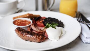 A plate of breakfast food including sausages, eggs, tomatoes, and toast, with a glass of orange juice in the background.